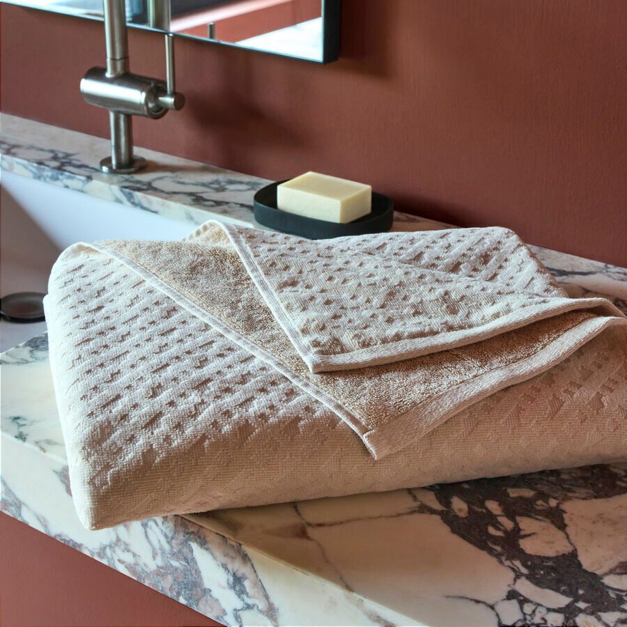Une serviette beige texturée, pliée, repose sur un meuble-lavabo en marbre, à côté d'un savon, avec un robinet en arrière-plan.