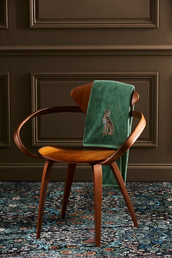 Chaise en bois au dossier incurvé, une serviette verte drapée dessus ; devant un mur sombre, sur un tapis à motifs.