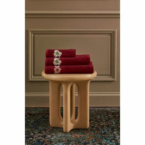 Pile de serviettes rouges à broderies florales sur un tabouret beige, devant un mur marron et un tapis à motifs.