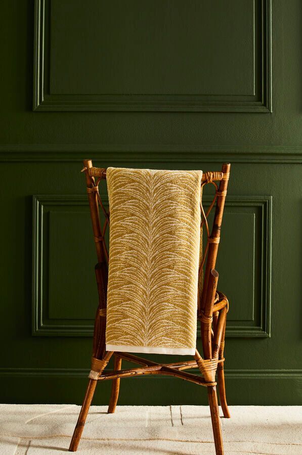 Une couverture dorée tissée est drapée sur le dossier d'une chaise en bambou brun, devant un mur lambrissé vert foncé.
