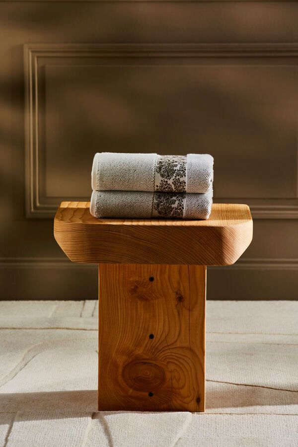 Deux serviettes grises à motifs sont empilées sur un tabouret en bois contre un mur marron.