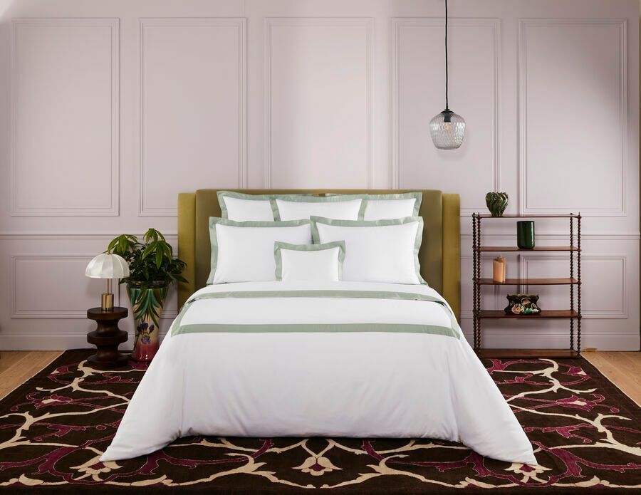 Draps de lit blancs à bordures vertes sur un lit dans une chambre avec un tapis marron et une tête de lit olive.