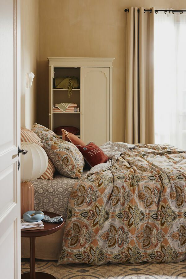 Chambre avec lit, linge de lit à motifs, armoire, table de chevet ronde et rideaux beiges.