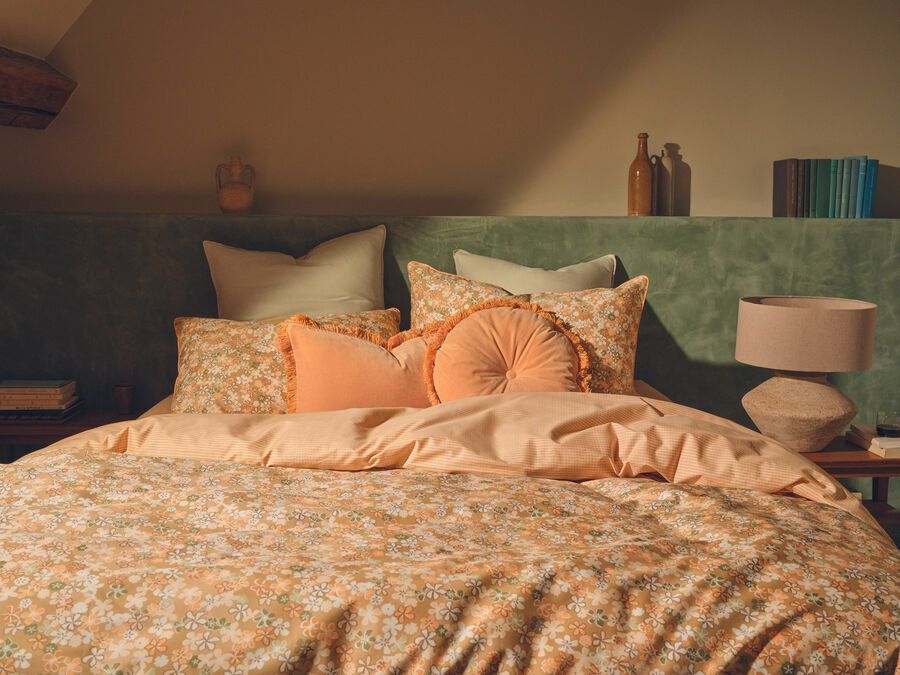 Un lit soigneusement fait avec des draps à motifs floraux, des coussins décoratifs couleur pêche et une tête de lit vert pâle dans une chambre à l'ambiance douce.