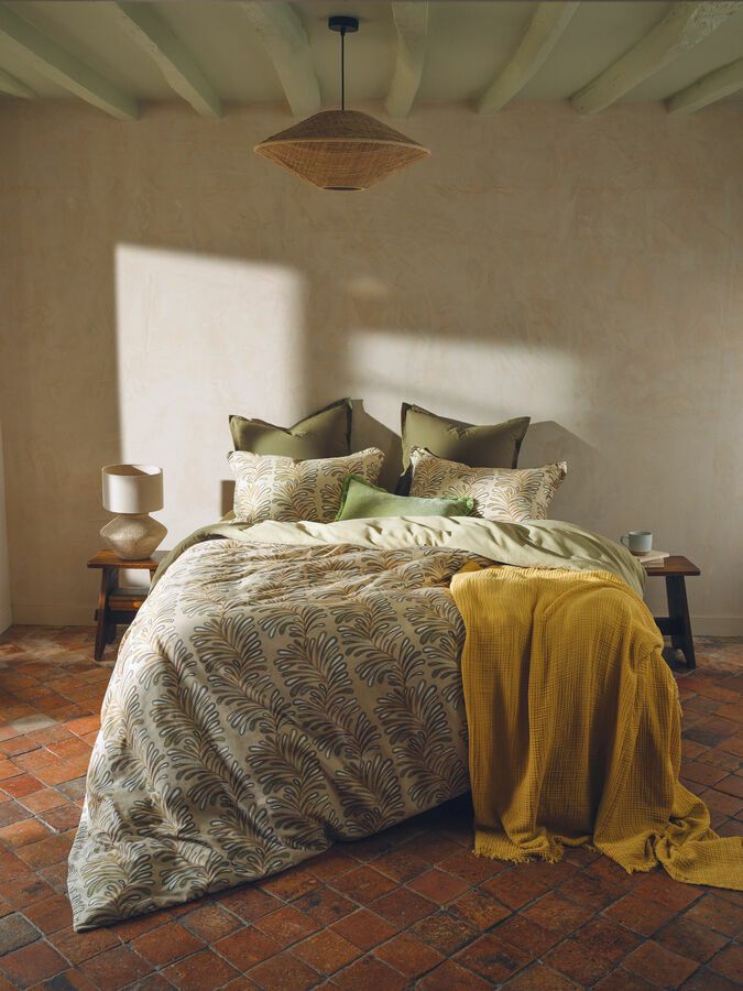 Une chambre avec un lit paré de draps olive à motifs, un jeté de lit moutarde et une suspension en rotin sur un sol carrelé.