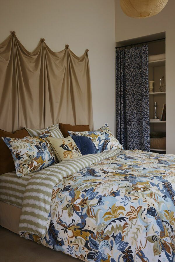 Chambre avec linge de lit à motifs floraux, tête de lit drapée beige, couette rayée. Rideau bleu marine à côté d'étagères encastrées.