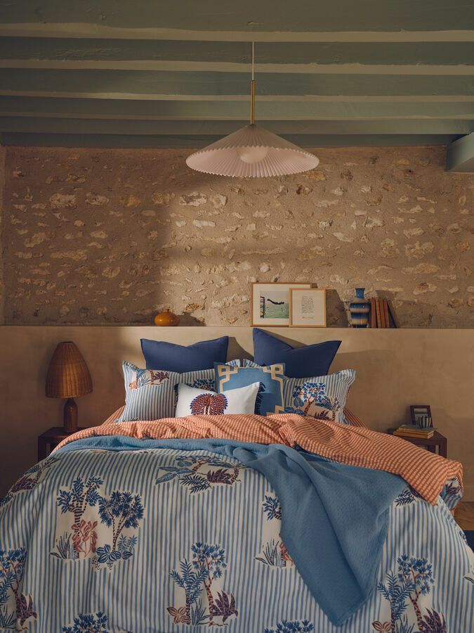 Un lit recouvert d'une couette bleue et blanche à motifs, surmontée d'un jeté orange, dans une chambre avec un mur en pierre et un plafond à poutres apparentes.