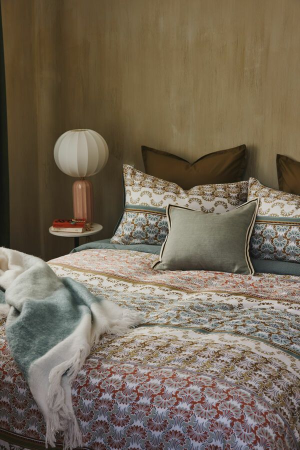 Chambre avec linge de lit à motifs, un jeté vert, une lampe ronde et un mur aux tons naturels.