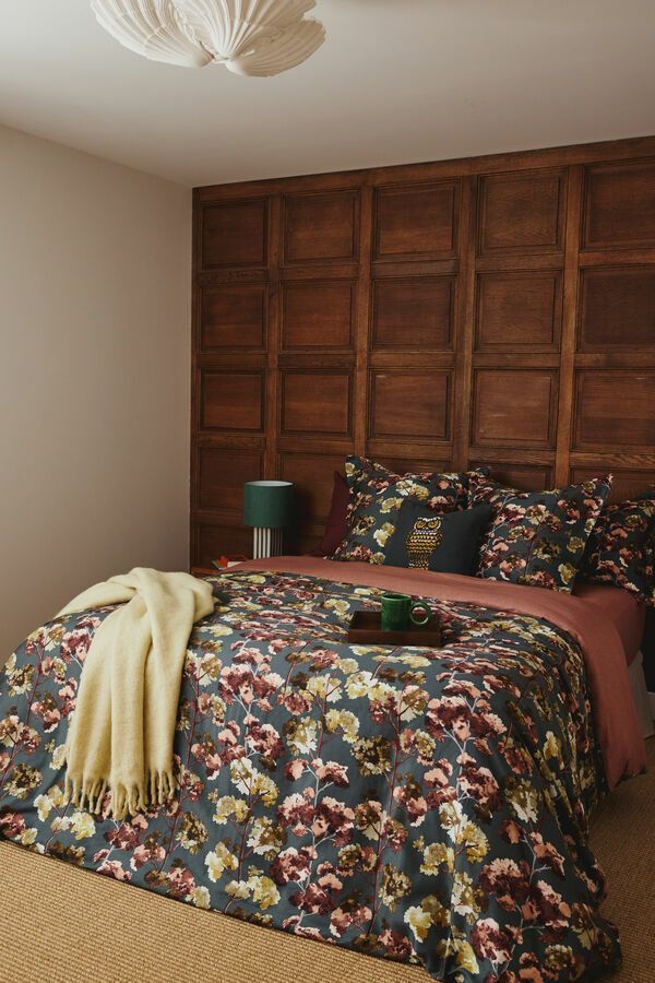 Chambre avec tête de lit en bois, linge de lit à motifs floraux et lampe verte sur la table de chevet.