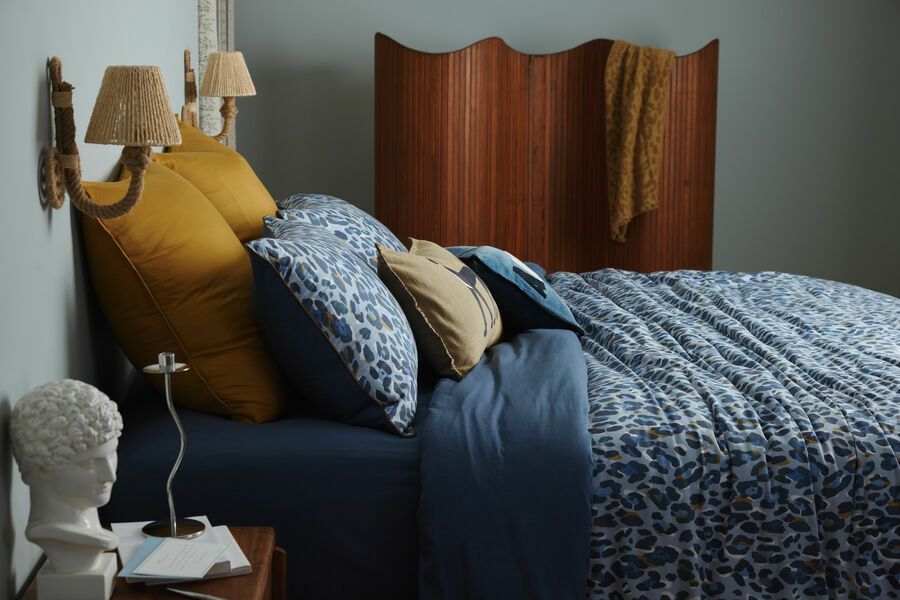 Un lit avec des draps bleus, des oreillers moutarde et une tête de lit en bois, avec une lampe et une sculpture sur une table de chevet.