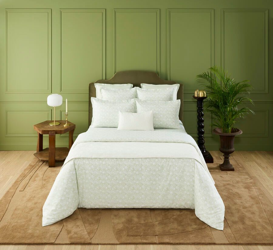 Un lit avec une literie à motifs clairs, placé au centre d'un mur lambrissé vert, avec une petite table en bois et une plante d'intérieur.