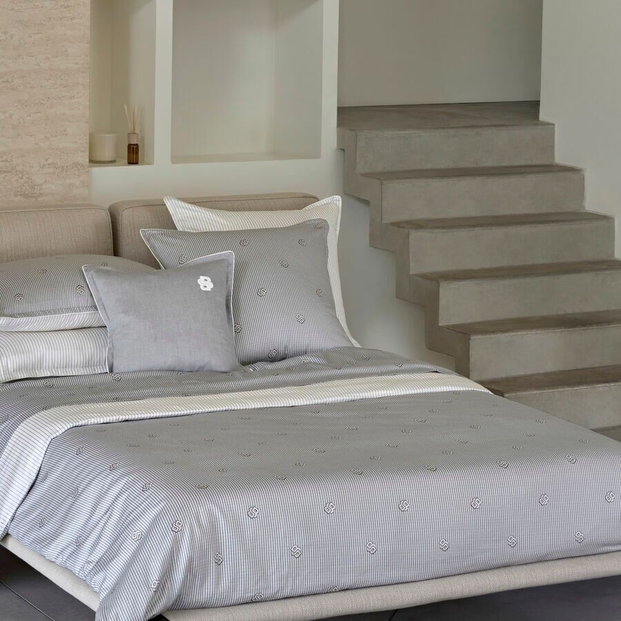 Un lit avec des draps gris à rayures et plusieurs oreillers, situé près d'une étagère ouverte et d'un escalier en béton.