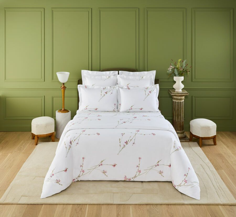 Un lit recouvert de draps blancs à motifs floraux est placé au centre d'un mur lambrissé vert olive, sur un parquet clair, avec deux tabourets.