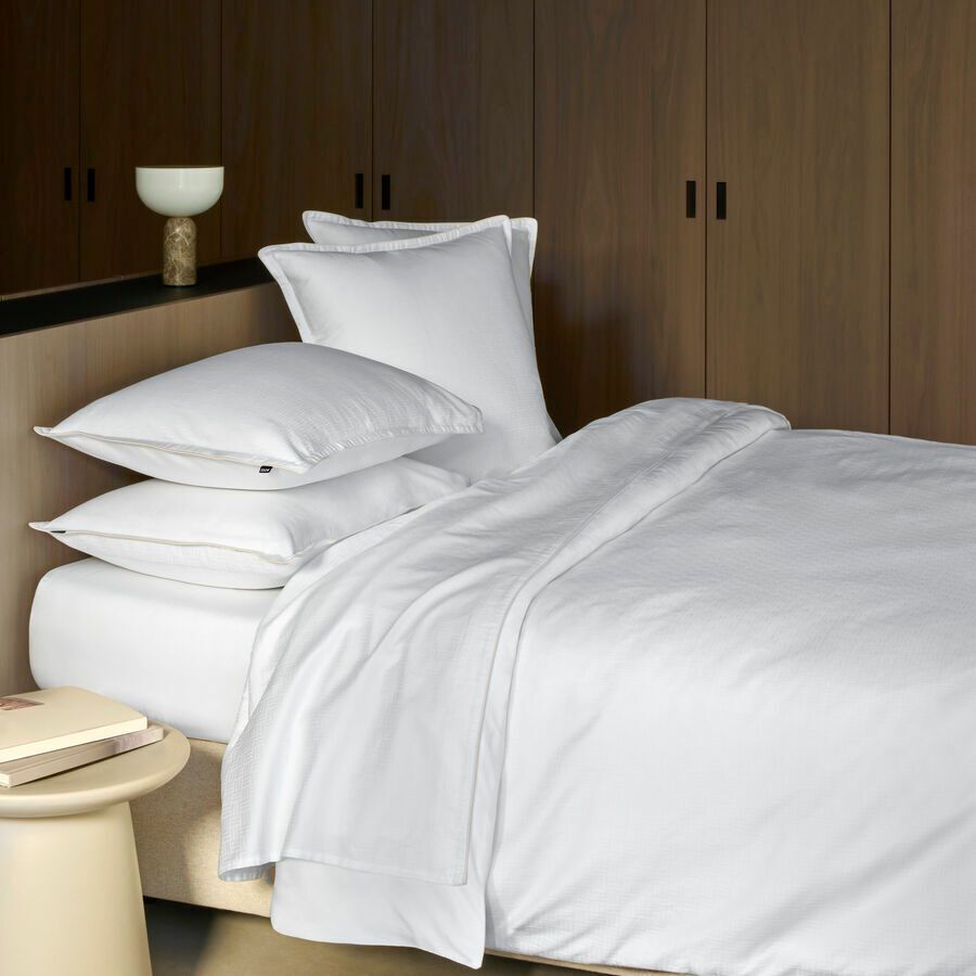 Un lit soigneusement fait avec des draps blancs, des oreillers et un plaid texturé, situé dans une chambre aux boiseries sombres.