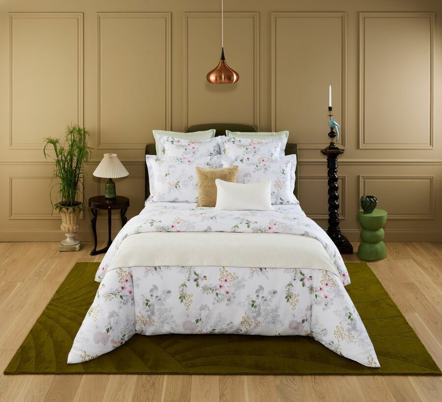 Un lit recouvert de draps à motifs floraux et d'un jeté couleur crème, adossé à un mur lambrissé, avec un tapis vert olive sur un parquet.