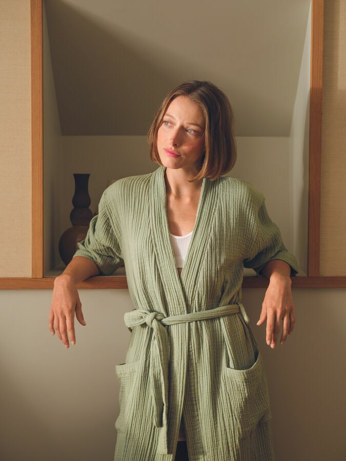 Une personne aux cheveux bruns mi-longs porte une robe texturée vert sauge et est appuyée contre une niche murale en retrait.