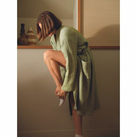 Une personne aux cheveux bruns mi-longs, vêtue d'une robe vert sauge, se tient debout tout en enfilant une chaussette claire.