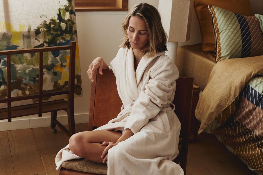 Une femme en robe de chambre blanche est assise sur un lit dans une chambre chaleureuse et ensoleillée.