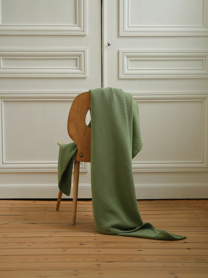 Un plaid texturé vert sauge drapé sur une chaise en bois, devant des portes à panneaux blancs sur un parquet.