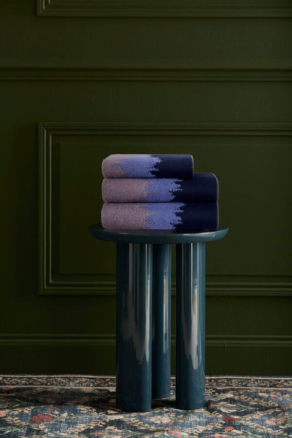 Pile de serviettes bleues et violettes à effet ombré sur une table trépied bleu sarcelle foncé, devant un mur vert.