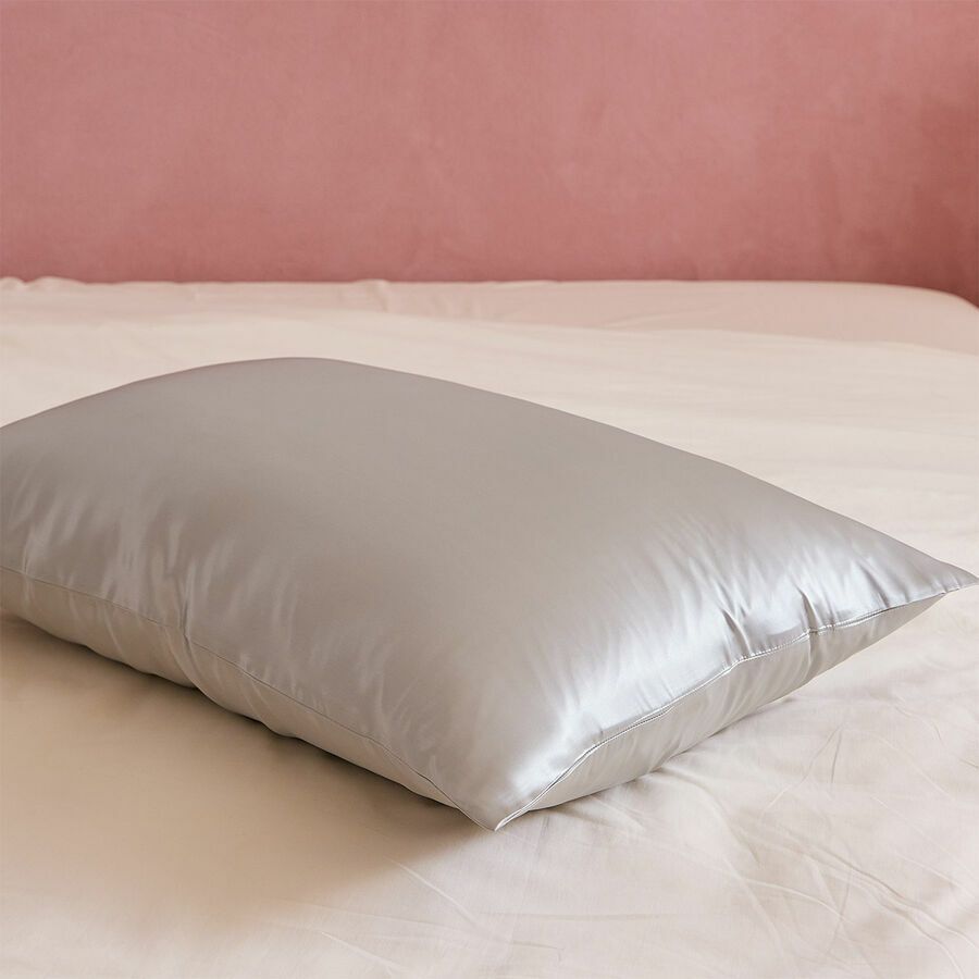 Un oreiller lisse en satin argenté repose au centre d'un drap blanc, devant un mur rose pâle.