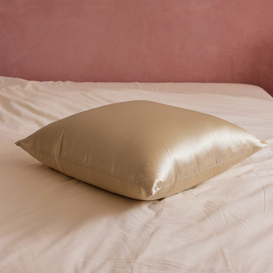 Un coussin carré brillant, couleur champagne, est posé sur un lit blanc, devant un mur rose pâle.