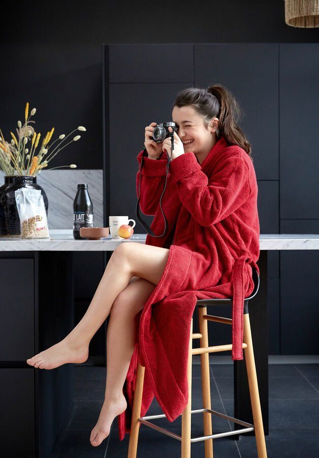 Une femme en robe rouge est assise sur un tabouret et prend une photo avec un appareil photo.