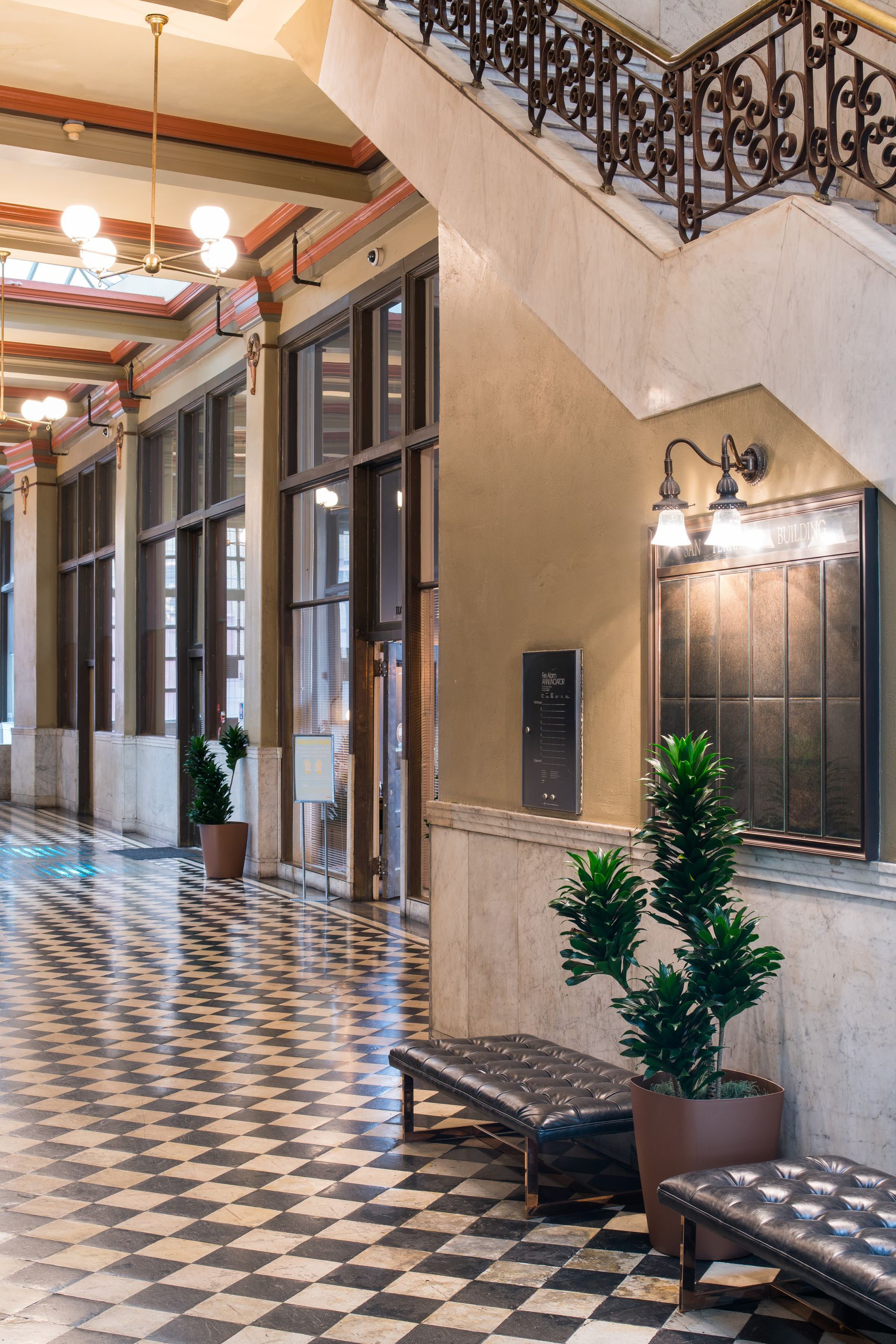 Elegant indoor hallway with checkered tile floor, tall windows, ornate railing, and potted plants.