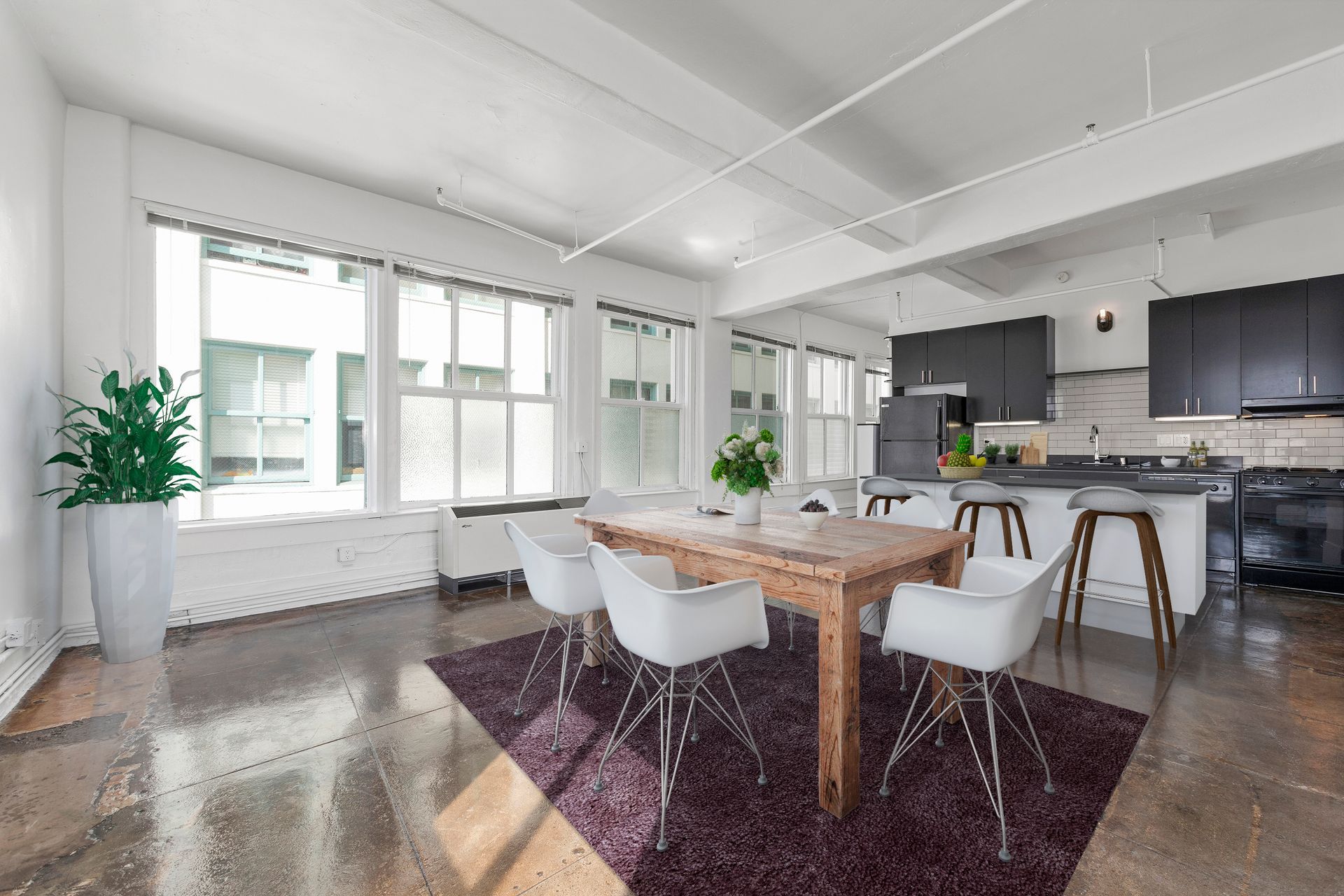 Bright open dining room with wooden table, white chairs, dark rug, and modern kitchen in the background
