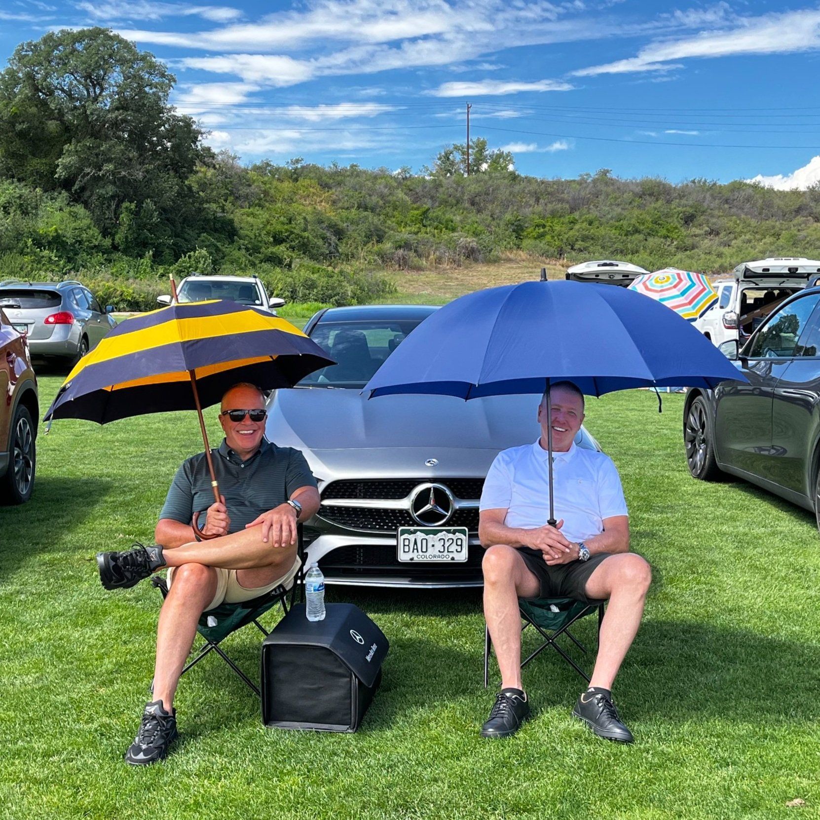 Two men are sitting under umbrellas in front of a car.