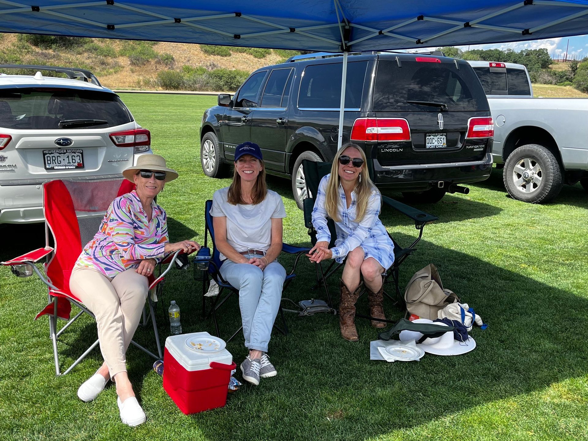 Three women are sitting under a blue tent in a field.