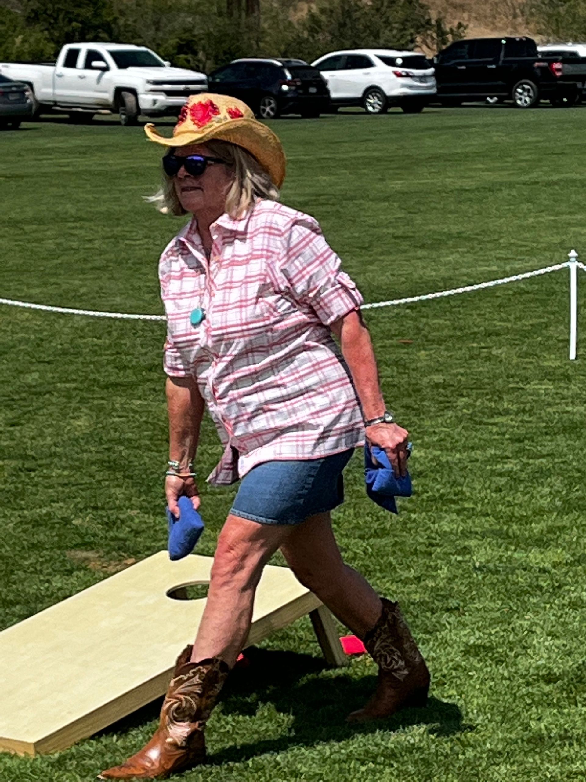 A woman wearing a cowboy hat and cowboy boots is playing cornhole in a field.