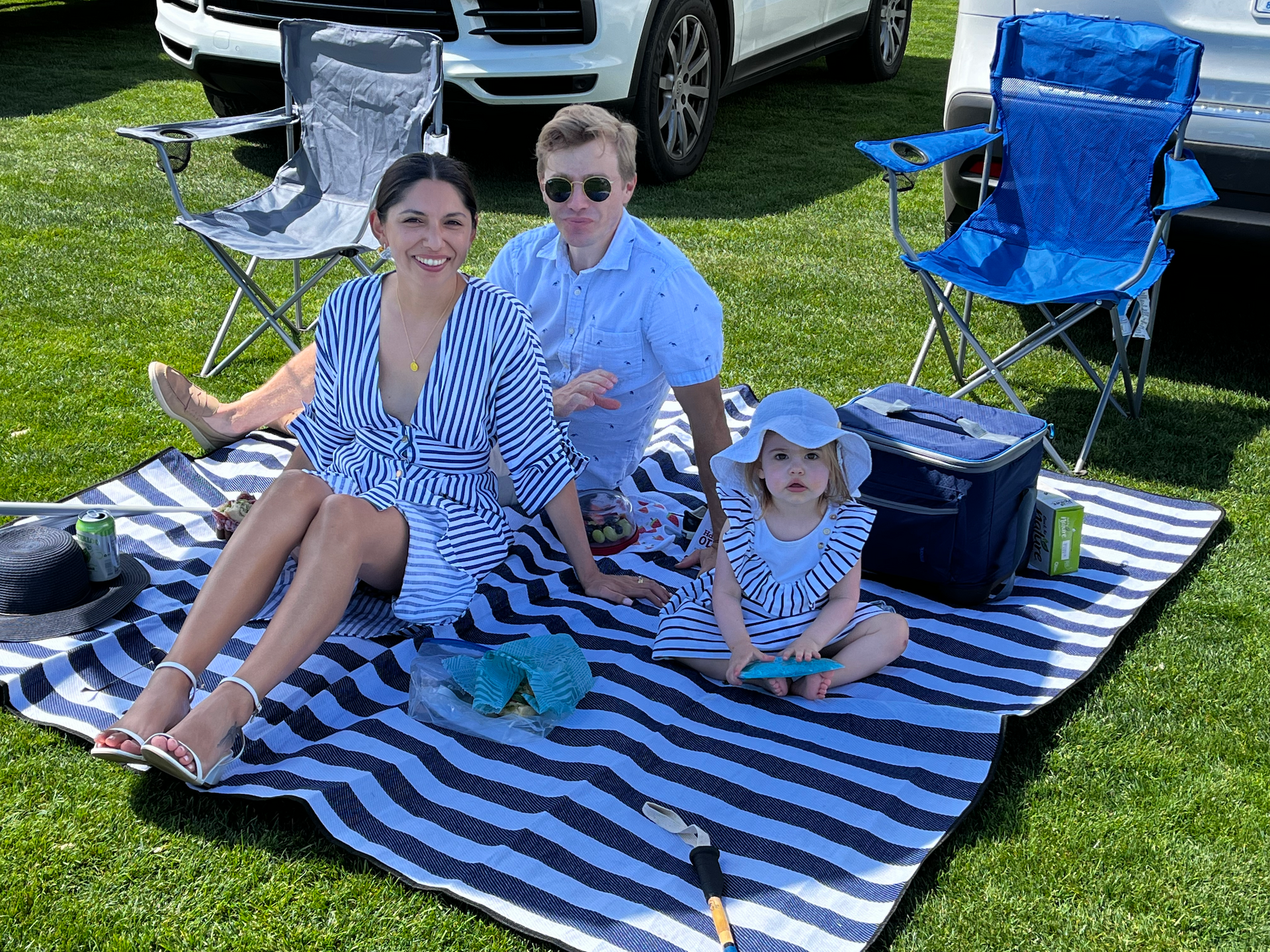 A family is sitting on a striped blanket in the grass.