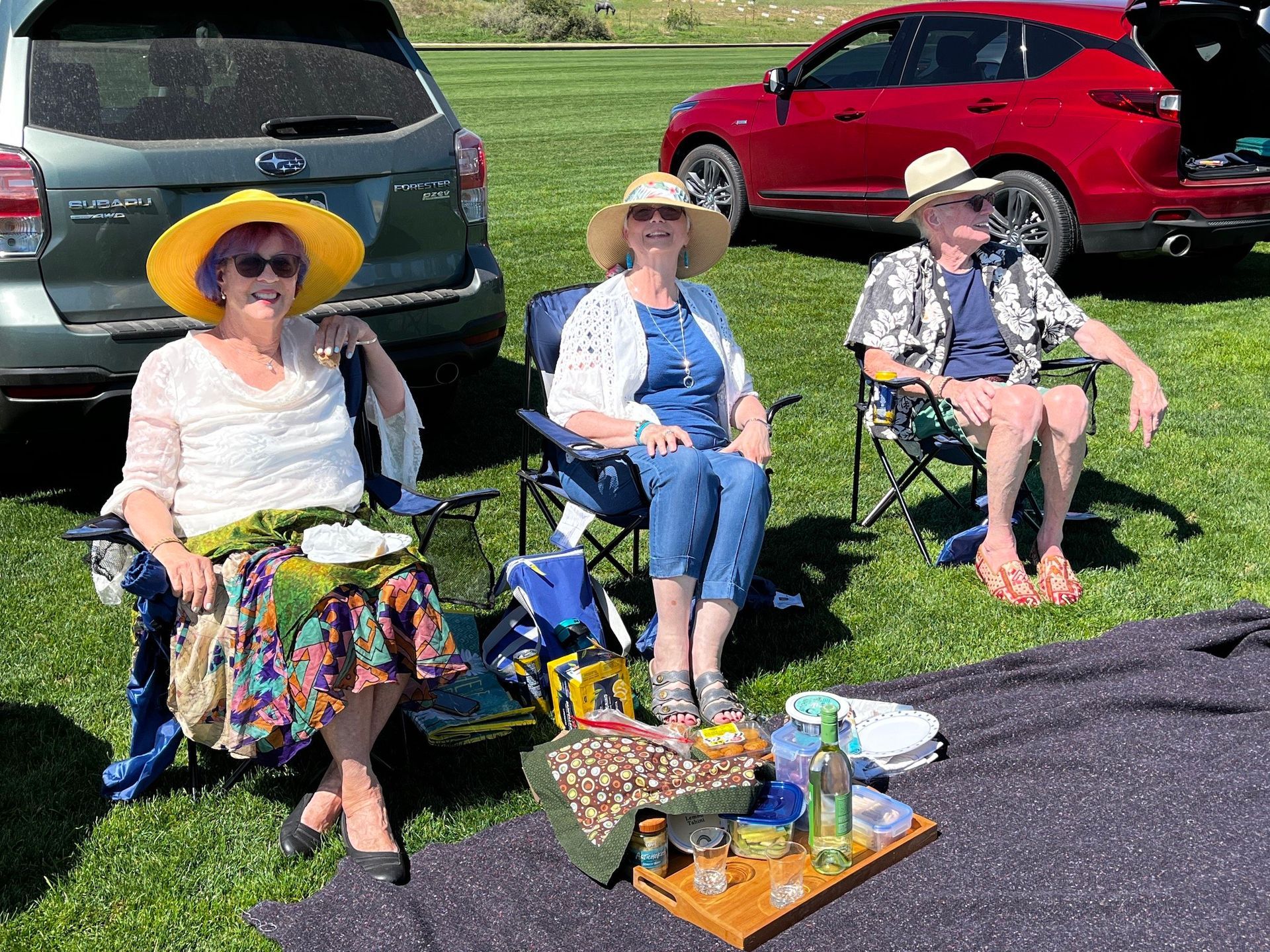 Three women are sitting in chairs in the grass in front of a red car.