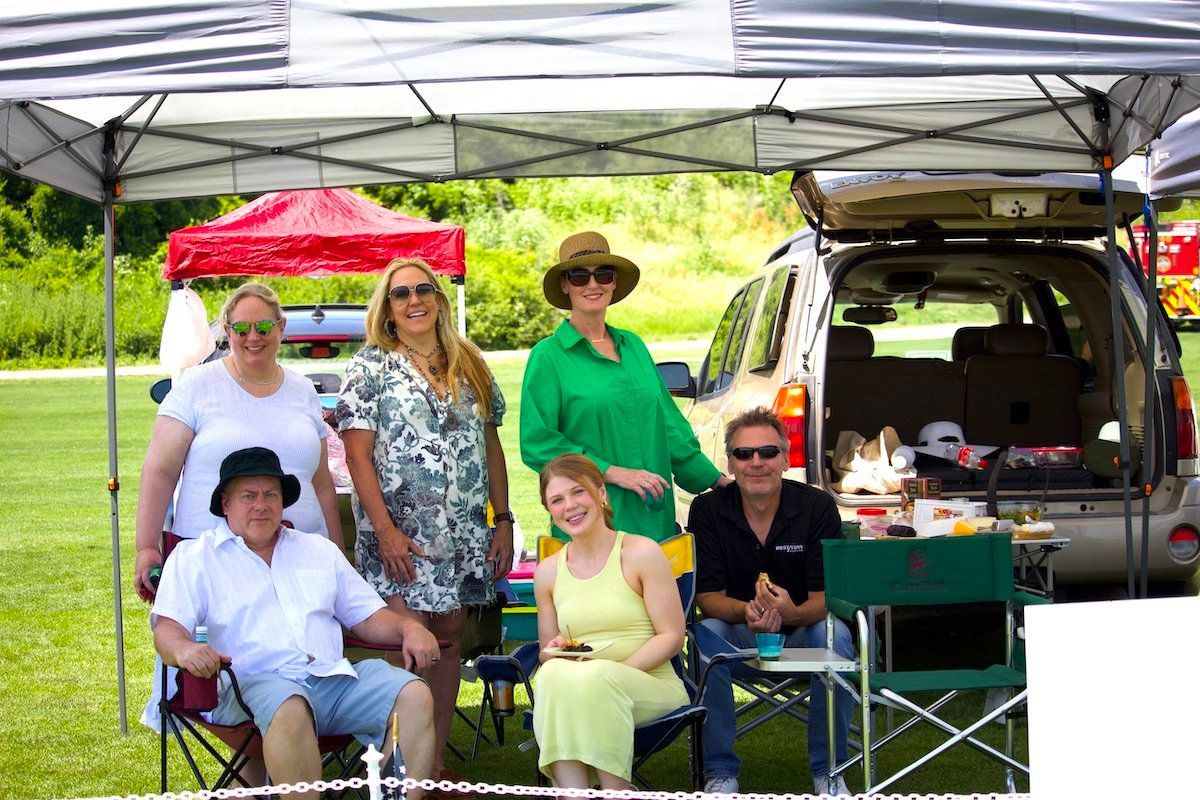 A group of people are sitting under a tent in front of a car.
