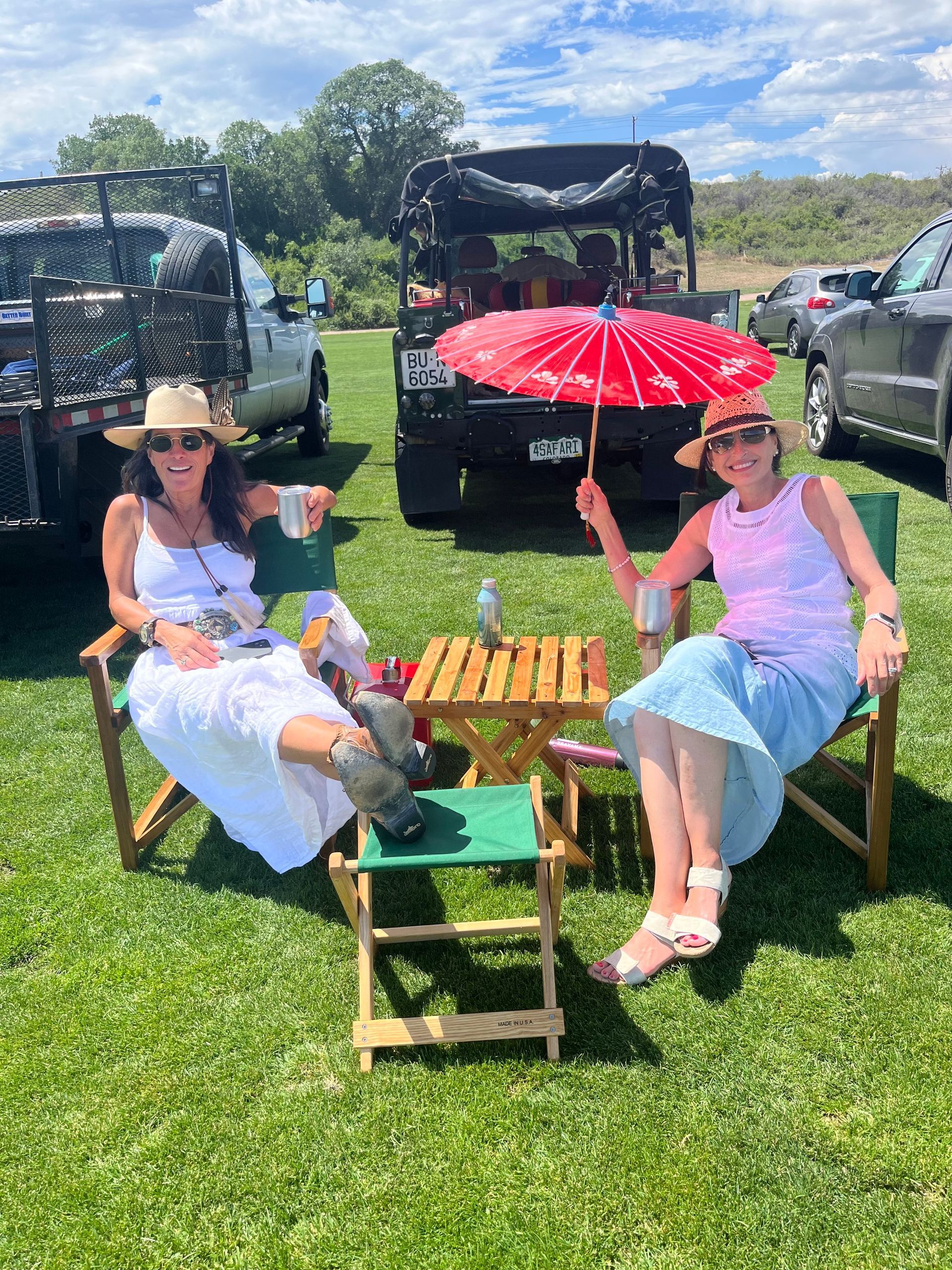 Two women are sitting in folding chairs in a field holding umbrellas.