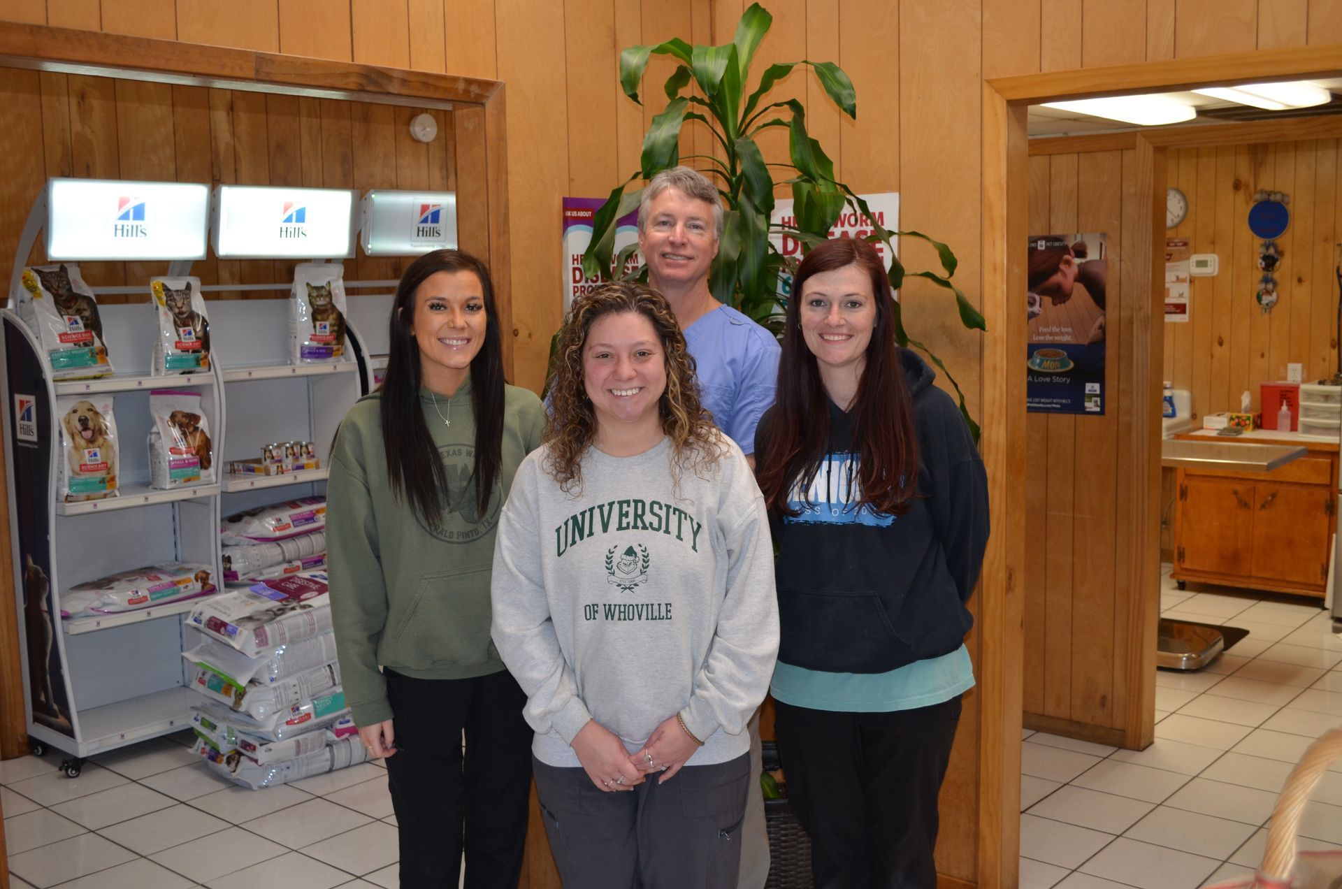 Four people in a veterinary clinic, smiling. A man in scrubs and three women stand near food displays and a plant.