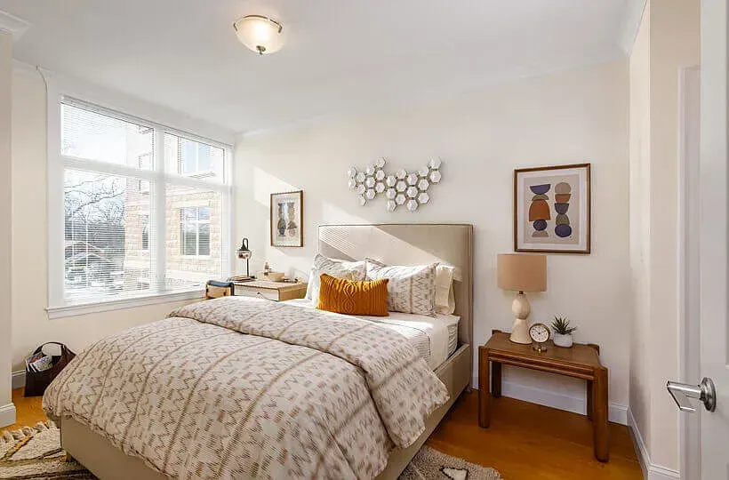 A sunlit bedroom features a bed with a neutral patterned duvet and mustard pillow, beside a wooden nightstand and window.