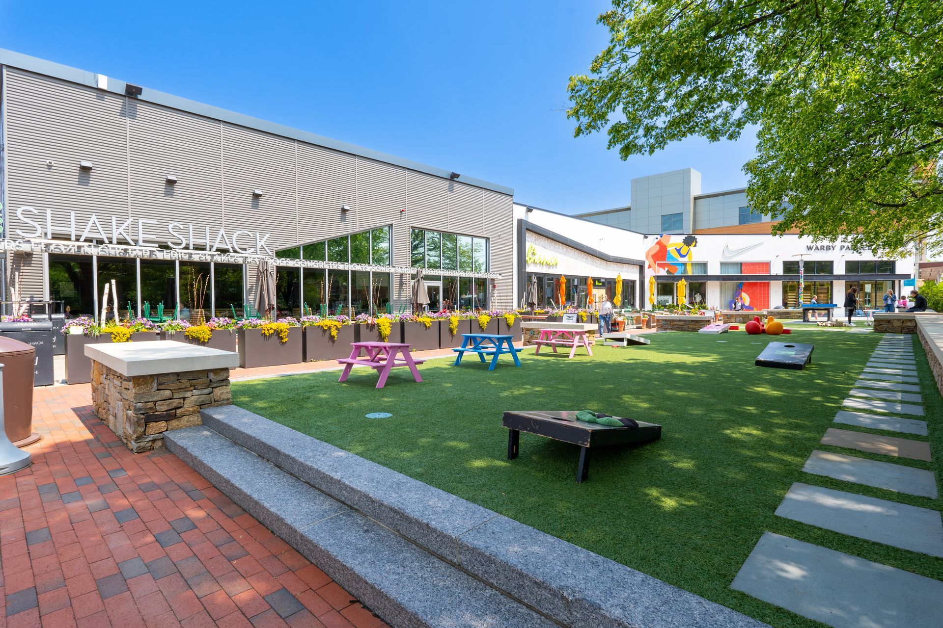 Outdoor plaza with colorful picnic tables on green grass, surrounded by buildings on a sunny day.