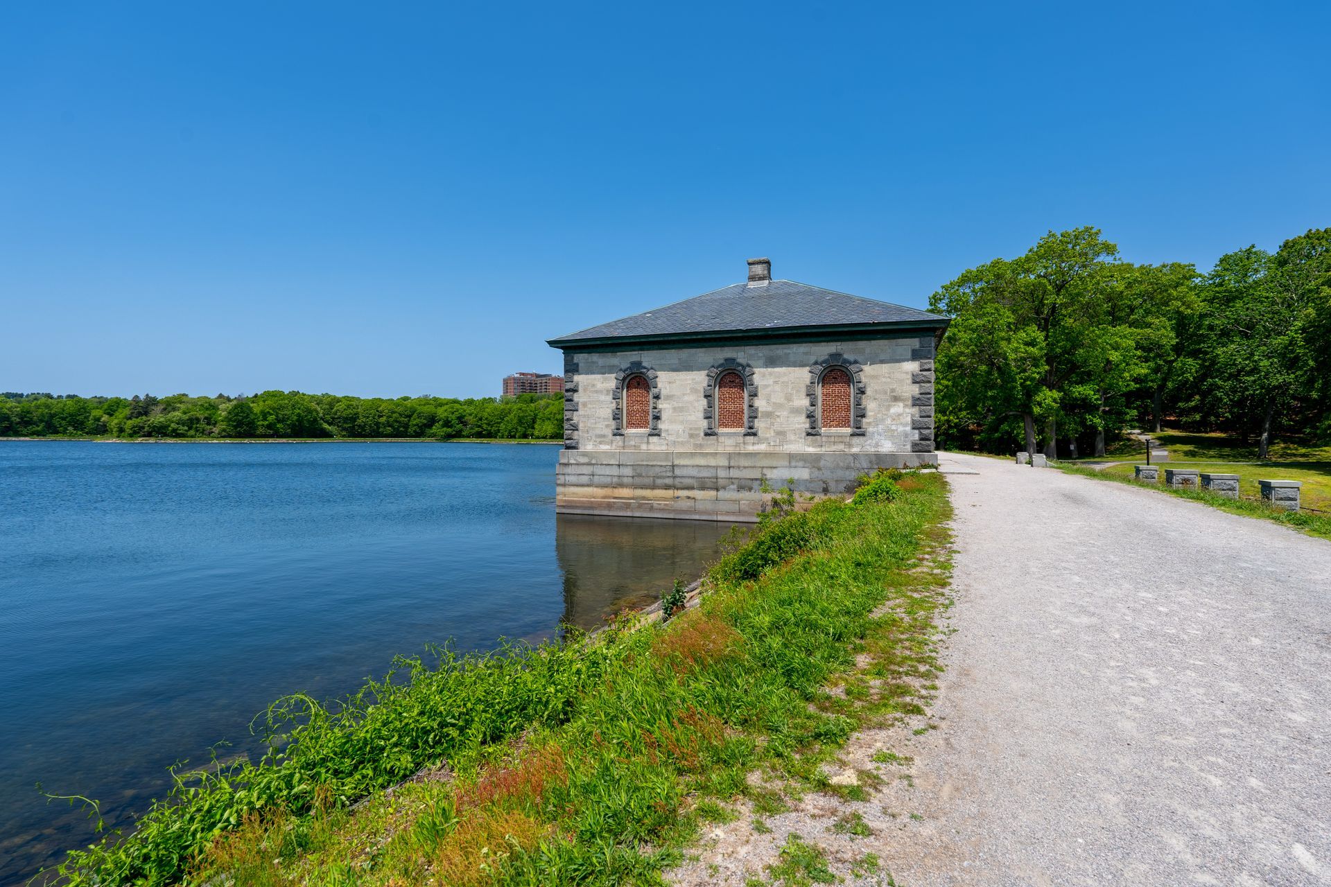 Stone building with dark roof by a lake, next to a gravel path. Green trees and blue sky.