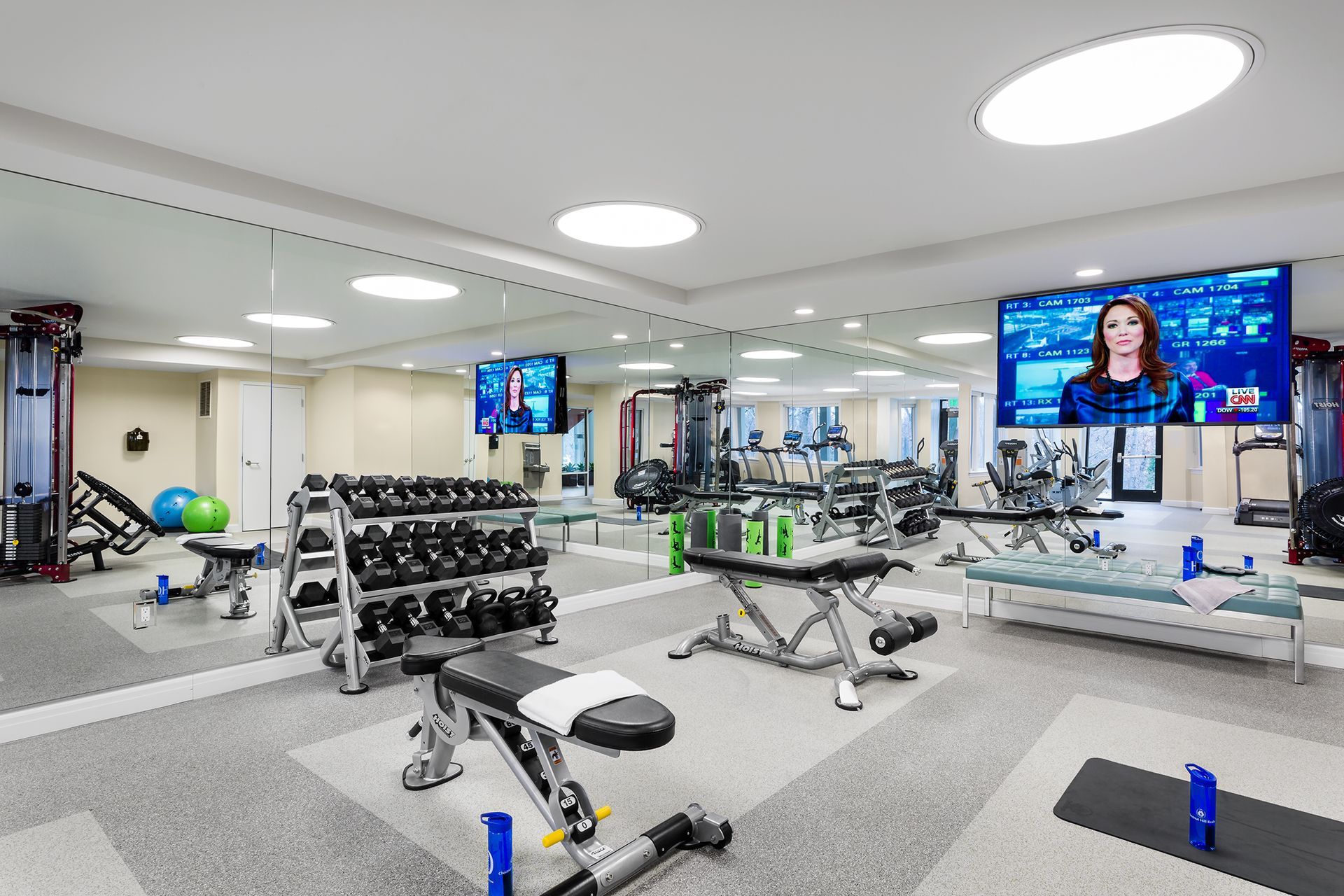 Gym interior with exercise equipment, including weights, benches, and machines, plus large mirrors and TVs.