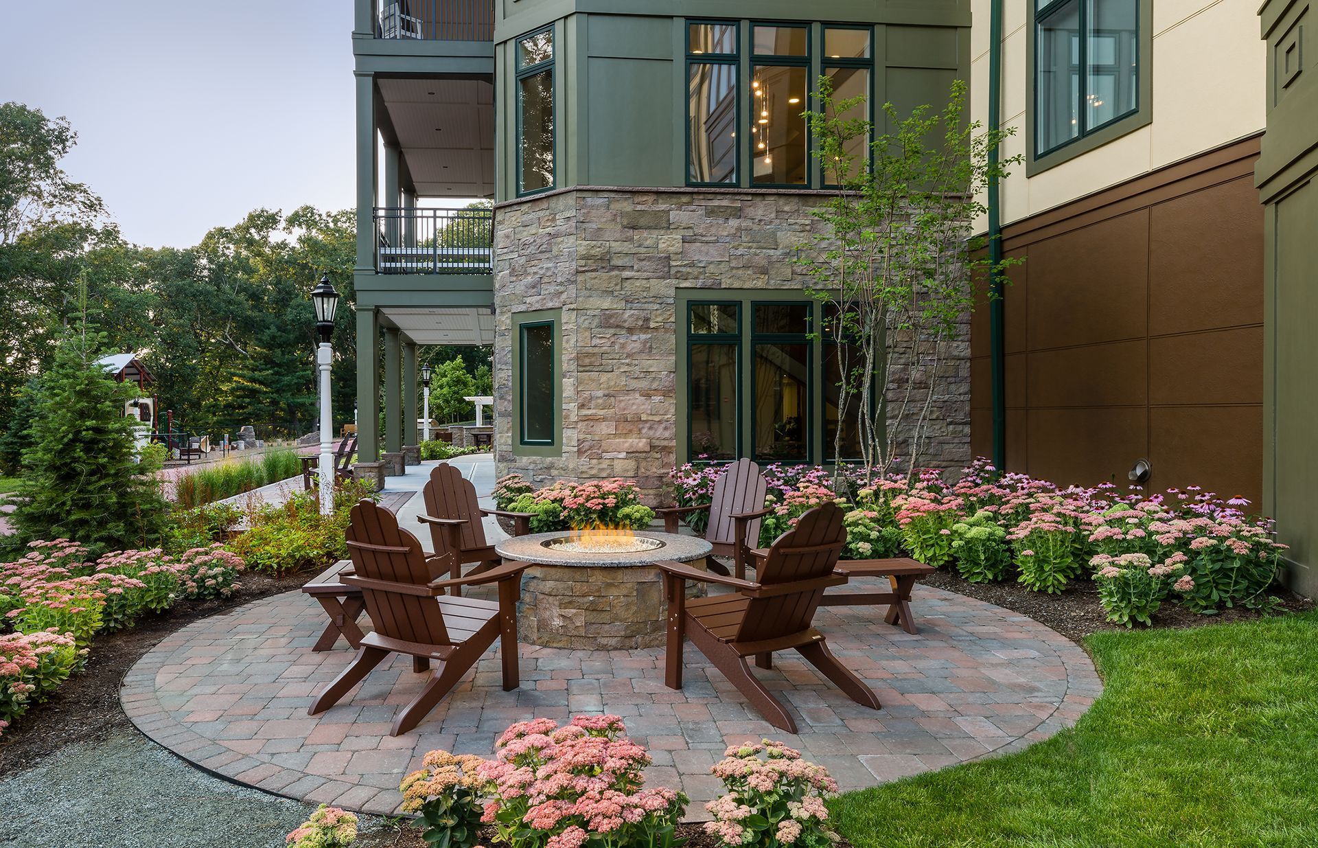 Stone patio with Adirondack chairs, fire pit, and flowering plants in front of a multi-level building.