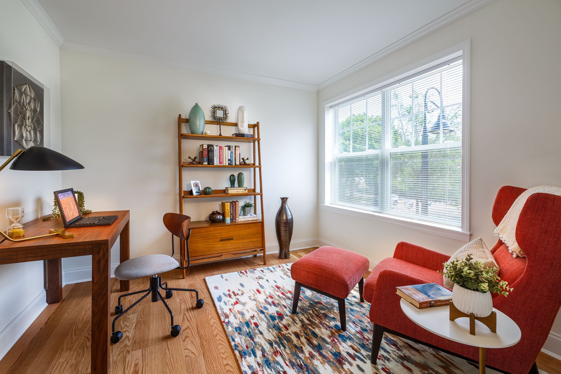 Home office with desk, bookshelf, red chair, and colorful rug. Window with blinds.