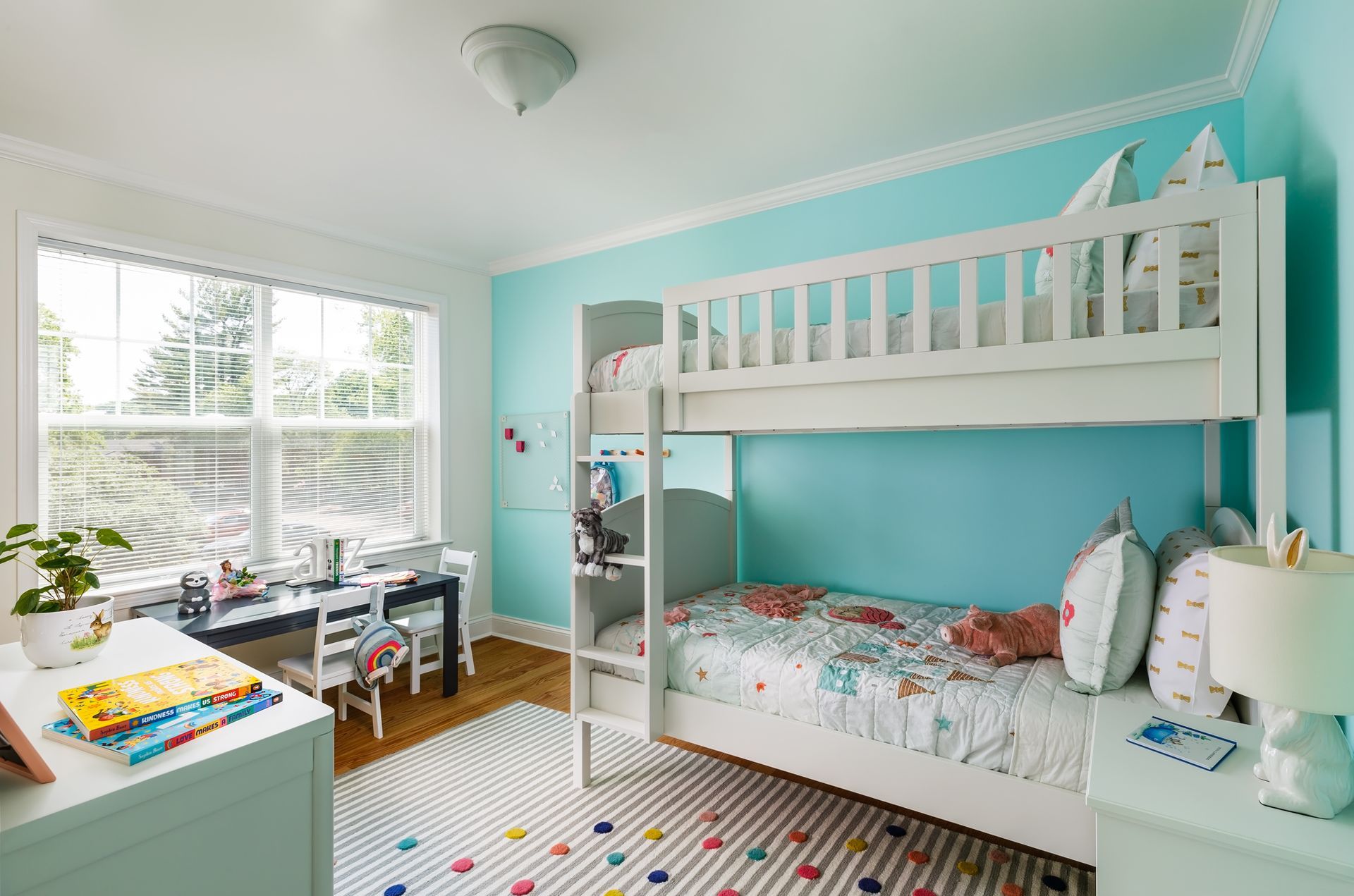 Children's bedroom with bunk beds, desk, and window. Turquoise walls, white trim, and a colorful rug.