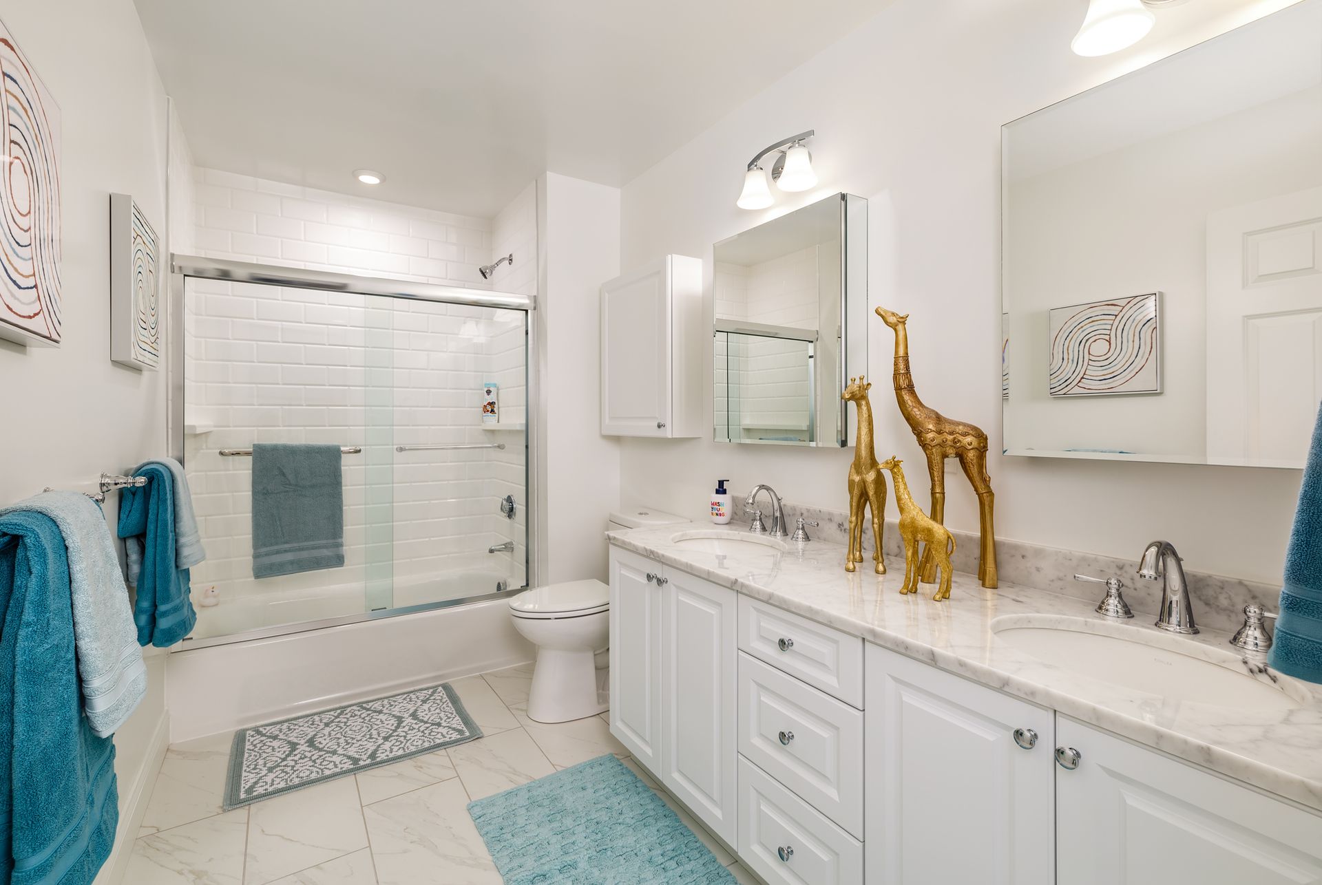 White bathroom with shower, vanity, and decorative giraffes. Teal towels and rug add color.