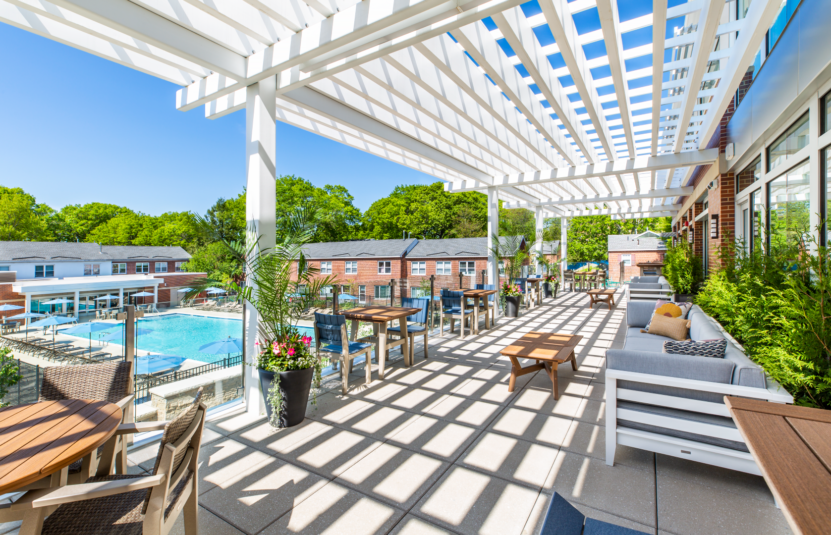 A large patio with tables and chairs under a pergola with a pool in the background.