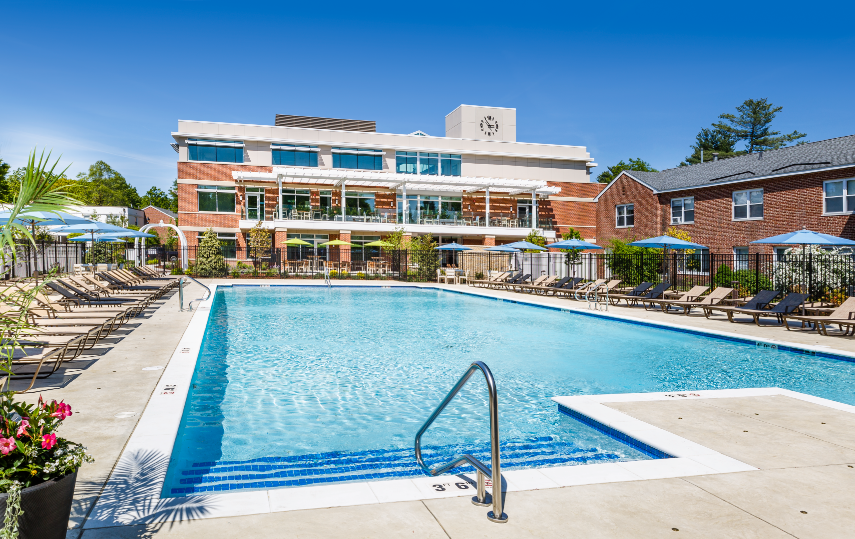 A large swimming pool surrounded by chairs and umbrellas in front of a brick building.