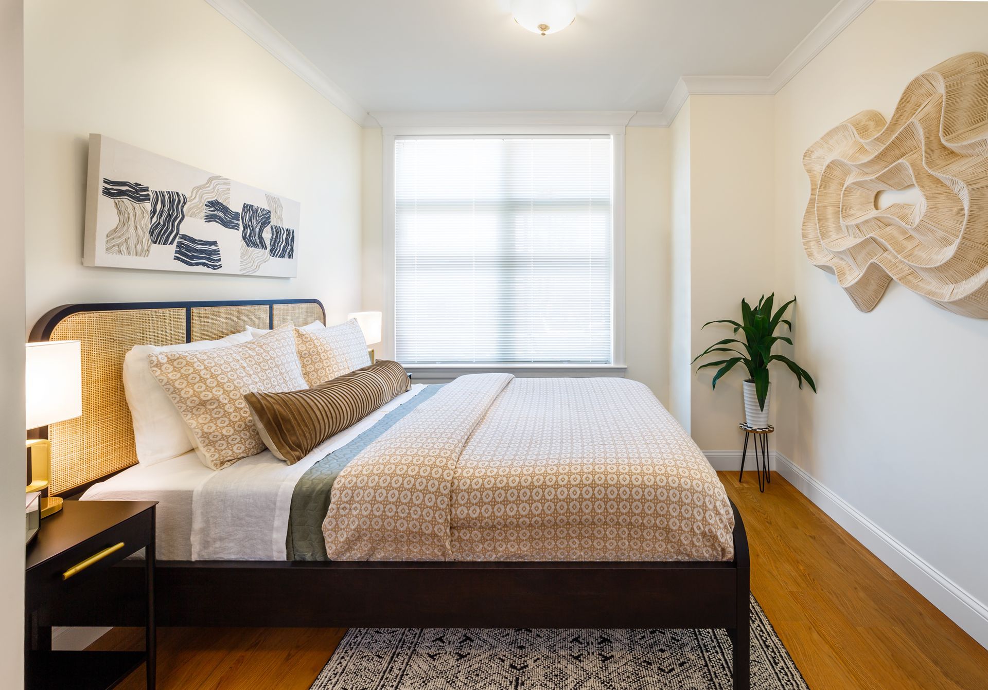 Bedroom with bed, art, small plant, wood floor, and window with blinds.