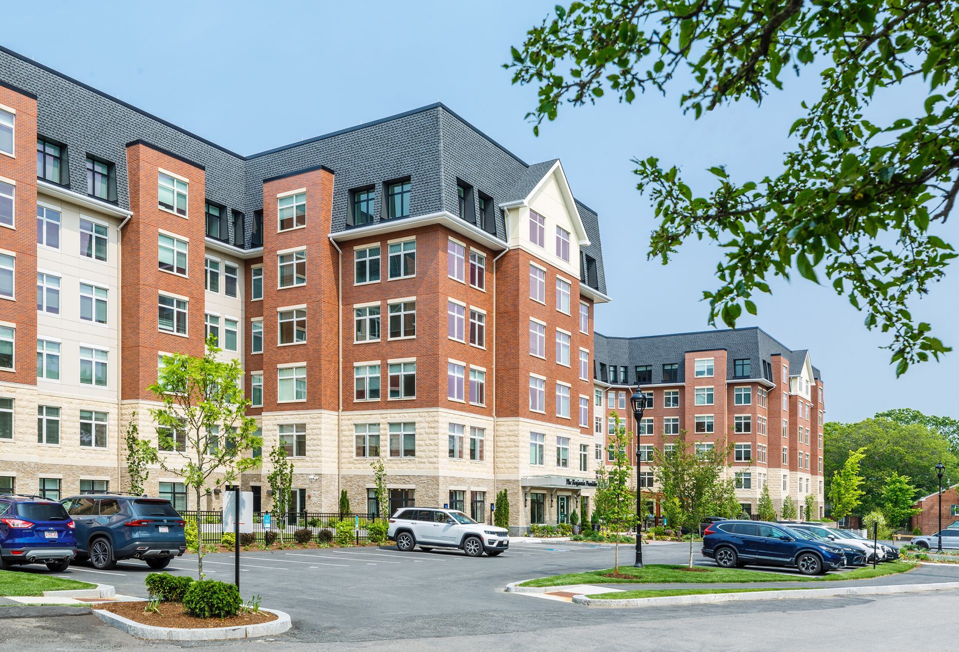 Multi-story brick apartment building with parked cars, blue sky, and greenery.
