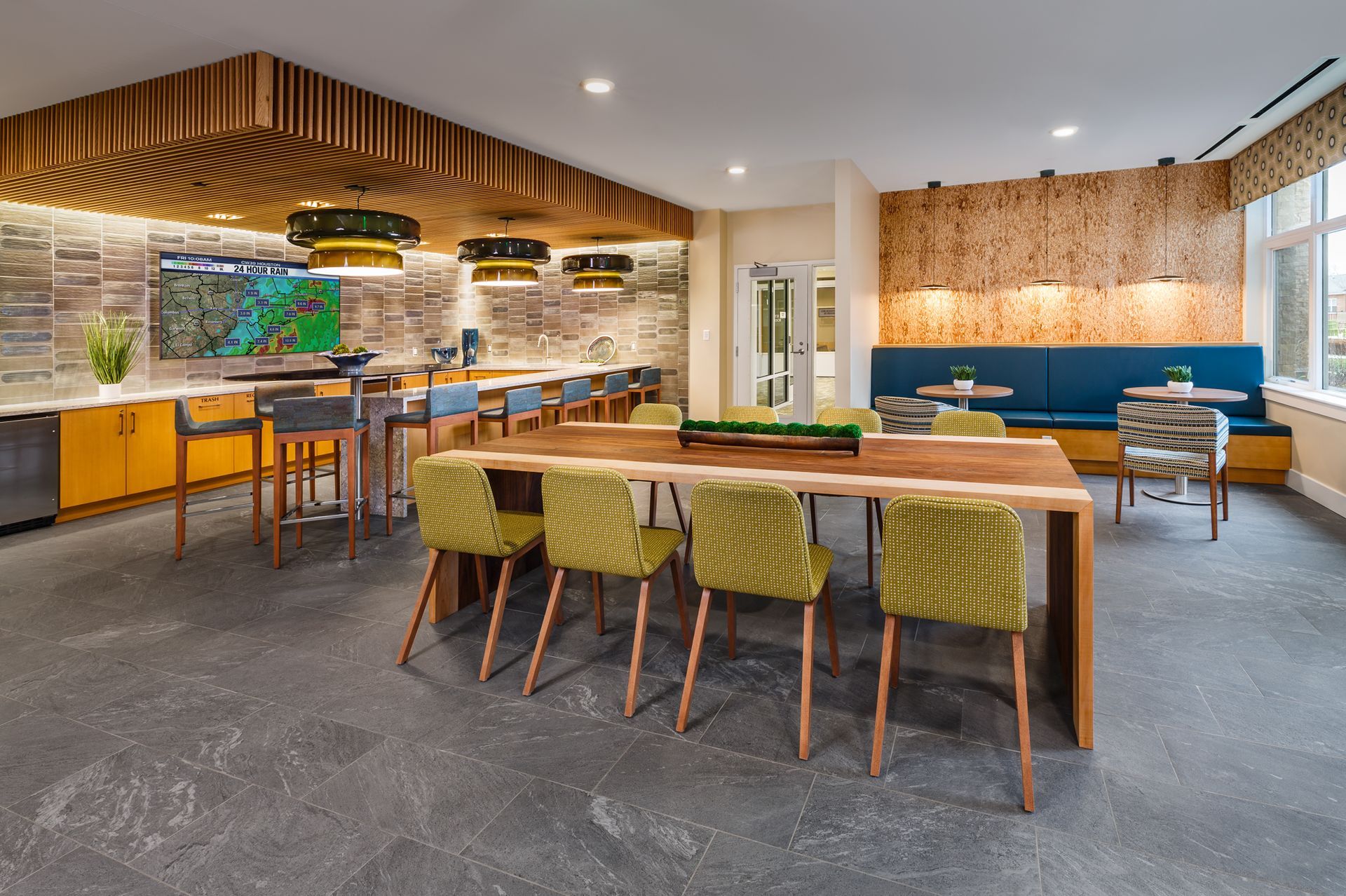 A modern lounge with a long wooden table, green chairs, and a bar area with stone and wood accents.